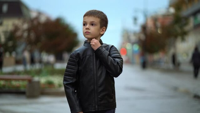 The child fastens the lock of the jacket on the city street. Young boy is zipping up jacket