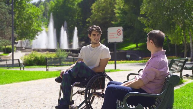 Disabled young man is chatting with his friend.
Disabled youth in wheelchair chatting with friend sitting on bench outdoors.

