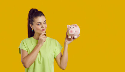 Studio shot of pretty woman holding piggy bank and thinking. Young girl considers investing but has some doubts. Beautiful lady wants to buy something, saves money and plans her expenses beforehand