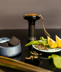 Black hookah bowl with aromatic tobacco with lemon and mint on black table with smoking accessories. Hookah smoking in a hookah lounge. Hookah bowl and coconut charcoal