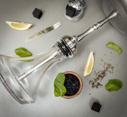 Hookah bowl with aromatic tobacco with lemon and mint on gray table with smoking accessories. Hookah smoking in a hookah lounge. Steel hookah, tongs, tea and coconut charcoal