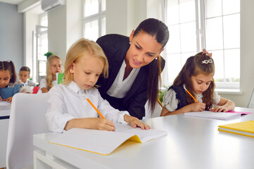 Friendly young female teacher helping little students with test tasks in primary school classroom. Teacher walks around classroom during lesson and gives hint to little schoolboy. Concept of education