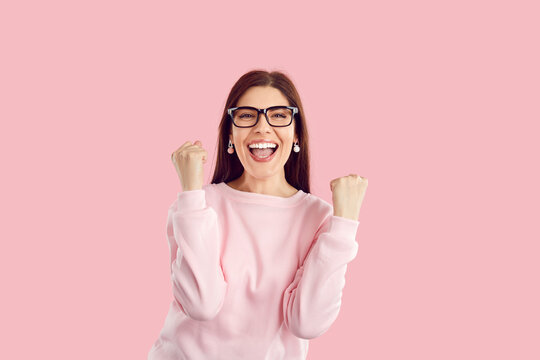 Happy excited woman rejoices success, clenched fists and triumphantly exclaims, isolated on pink background. Beautiful caucasian young woman in pink sweater and glasses achieved her aim and goals.