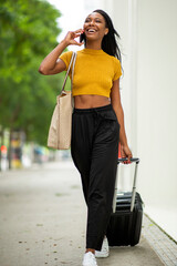 Happy black woman talking on smart phone while carrying luggage on city street