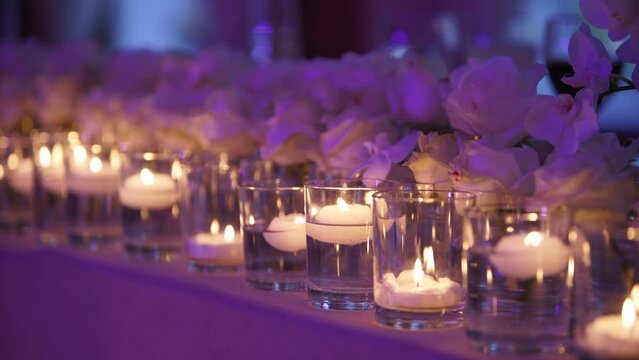 Candles floating in glass vases flasks filled with water. Banquet hall interior floristic details. Wedding decor with white roses on served newlyweds dining table setting at restaurant. Party lights.
