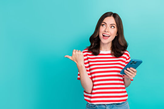 Photo of impressed dreamy lady wear striped t-shirt typing modern device pointing thumb empty space isolated turquoise color background