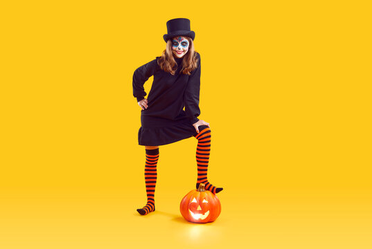 Full body shot of child in Halloween costume. Happy girl with skull makeup, wearing black dress, top hat and striped stockings steps on orange jack o lantern while posing on yellow studio background