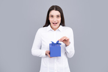 Surprised woman holding gift. Smiling beautiful young brunette woman in shirt hold present box with gift ribbon bow isolated on bright gray background, studio portrait. Birthday concept.