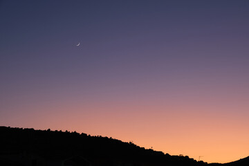 View of a crescent moon and mountain in silhouette at sunset.