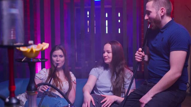 a company of young people, a guy and two young girls girlfriends sit relaxed in a hookah lounge, drink tea and smoke a hookah and chat merrily