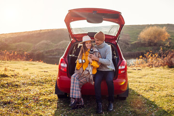 Young couple in love relaxing in car trunk by autumn lake drinking tea from vacuum flask. Man and woman travel by auto. - obrazy, fototapety, plakaty