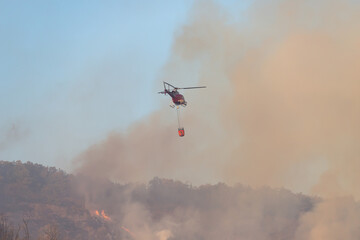 A fire helicopter carries water to extinguish a fire on Mount San Cristóbal, Pamplona. Heavy dark smoke covers the sky.