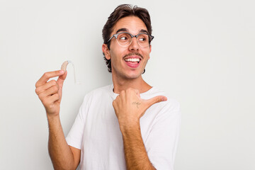 Young hispanic man holding hearing aid isolated on white background points with thumb finger away,...