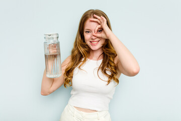 Young caucasian woman holding jar of water isolated on blue background excited keeping ok gesture on eye.