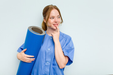 Young caucasian physiotherapist holding a mat isolated on blue background relaxed thinking about...