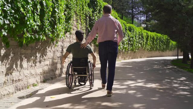 The young man in the wheelchair walking in the spring park and the mature man accompanying him.
He is accompanied by his physically disabled young friend walking on the street.
