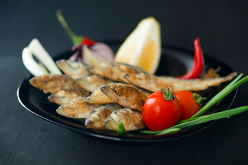 Small fried herrings with vegetables on a black plate on a black background. Fish appetizer.