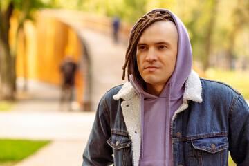 Fototapeta premium Portrait of handsome man with brown dreadlocks in hoodie, denim jacket posing on street in city park.