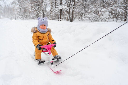 Sad toddler rides through the snow in a winter park on a children's snowcat holding on to the pink steering wheel