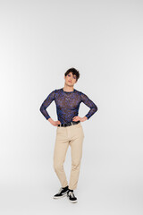 full length of nonbinary model in beige trousers posing with hands on waist on grey background.