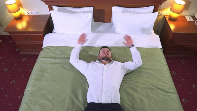 Tired male businessman in a white shirt and trousers falls on the bed after a hard day's work. Rest in a hotel room. Top view.
