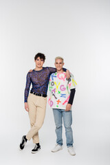 full length of young and joyful queer person with gay man hugging and looking at camera on grey...