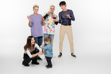 happy lgbtq friends blowing soap bubbles near toddler boy on grey background.