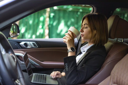 Successful attractive business woman sitting in the passenger seat in her luxury car working on laptop, holding a hot coffee mug and driving to a business meeting