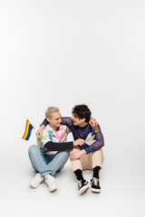 happy gay man and nonbinary person with small lgbtq flags embracing while sitting on grey background.