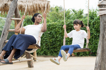 Happy young African American woman and child asian girl playing on swing together at the playground