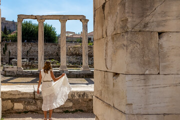 Rear view of tourist woman wearing white dress and golden laurel crown walking through ruined...