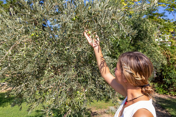 Woman wearing white dress and golden laurel crown holding a branch of olive trees in the garden of...