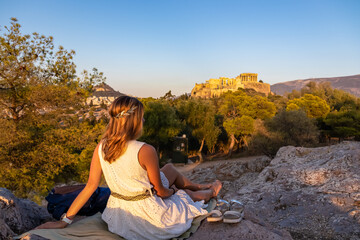 Naklejka na ściany i meble Rear view of tourist woman wearing a white dress and golden laurel crown looking at Parthenon of the Acropolis of Athens during sunset seen from Pnyx Hill, Attica, Greece, Europe. Ruins ancient temple