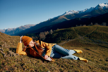 Fototapeta premium Woman sitting on a hill on the grass rest smile with teeth looking at the mountains in the snow in the winter in a yellow raincoat and jeans happy sunset trip on a hike, freedom lifestyle 