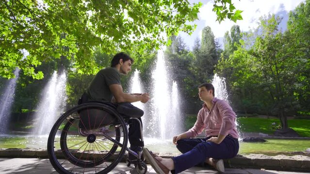 Young disabled man sitting in a wheelchair talking to his friend outdoors.
The physically disabled young man is having a conversation with his friend in the city park.

