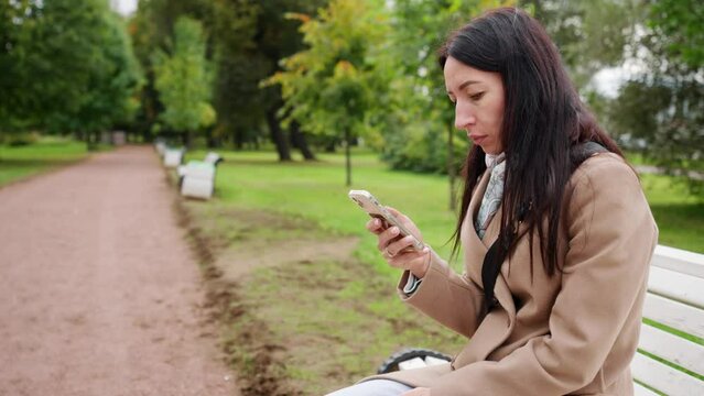 A young woman sits on a bench with a phone in her hands. In the background green park.