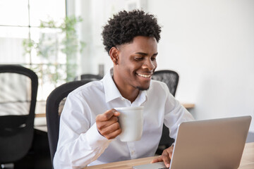 Happy male office worker with cup using laptop. Smiling businessman sitting at desk, drinking coffee while typing on computer. Relaxing, office work concept.