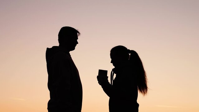 Silhouette man gives gift to his beloved woman outdoors against sky. Young guy gives box with surprise to his girlfriend, happy couple hugs, gentle kiss in nature. Valentines Day. Romantic love, park