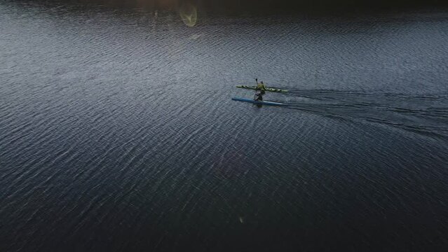 Aerial view on two unrecognizable people swimming on the kayak on the lake at sunset, following drone shot