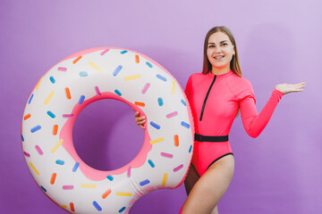 Slim young woman in a stylish pink swimsuit with a donut inflatable ring on a plain background.