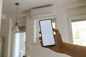 Close-up view of a man holding smartphone with blank screen ready for mock up. Adjusting his home air conditioner with a smart phone app. Smart home mobile phone control.