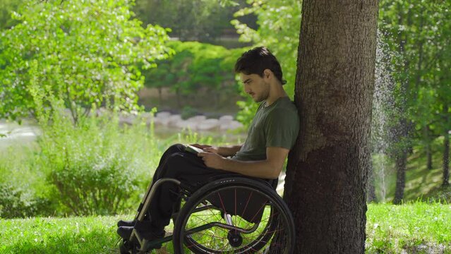 Teenager sitting in a wheelchair is reading a book.
Young man with physical disability reading a book outdoors.
