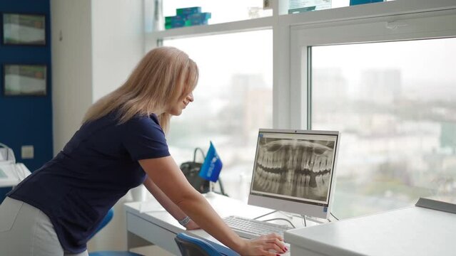 Beautiful Woman Dentist Examines a Picture of a Human Jaw on a Computer Monitor Screen. Woman Professional Orthodontist Smiles at the Camera. Concept of Dental Treatment and Medicine. Slow Motion
