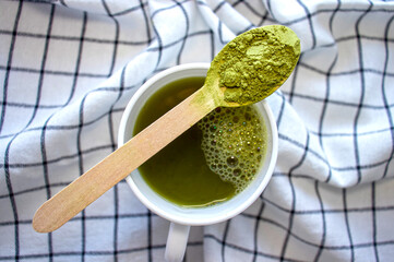 Matcha green tea in a white cup with powder in a wooden spoon against a checkered kitchen towel