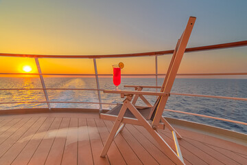 Fototapeta premium Vue d'une chaise longue avec un cocktail sur le pont d'un navire de croisière au coucher de soleil dans le sillage du navire de croisière. Vue depuis la poupe du navire.