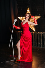Sexy young girl singer sings into a silver retro microphone in a red evening dress. Dark background in the photo studio
