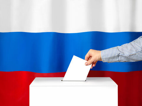 Voter's Hand Drops The Ballot Into The Ballot Box. Against The Background Of The Russian Flag. Filling Out Ballots And A Referendum In Ukraine. The Concept Of Elections