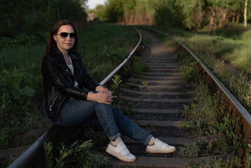 girl on railway, young woman in sunglasses and black leather jacket sits on railroad tracks. Brunette sitting on rail far.