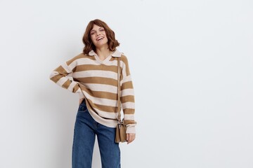 a pretty sweet woman is standing on a light background in a striped sweater and a bag on her shoulder standing half sideways with one hand on her hip lifting the sweater and lowering the other hand