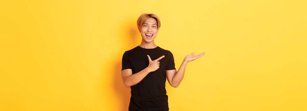 Portrait Of Handsome Smiling Asian Guy Holding Something On Hand And Pointing Finger At It, Standing Yellow Background Pleased